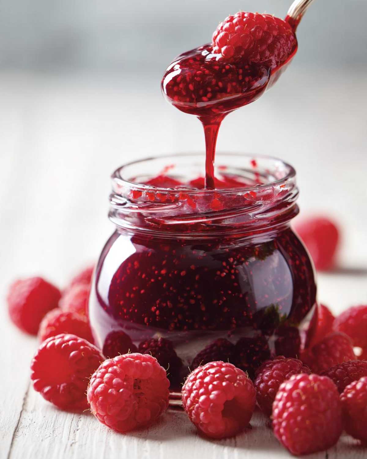 A spoonful of thick, glistening raspberry jam is dripping back into a glass jar surrounded by fresh, bright red raspberries.