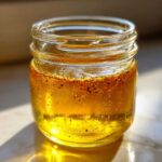 Close-up of golden homemade salad dressing separating in a small glass jar, showing spices settled at the top.