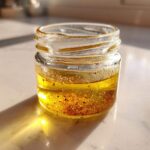 A small glass jar containing homemade salad dressing, featuring separated oil and spices, catching bright sunlight.
