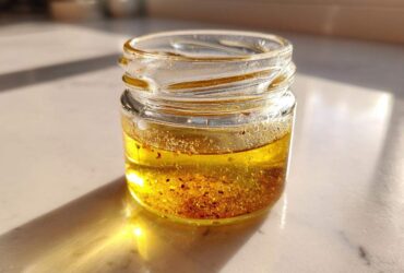 A small glass jar containing homemade salad dressing, featuring separated oil and spices, catching bright sunlight.