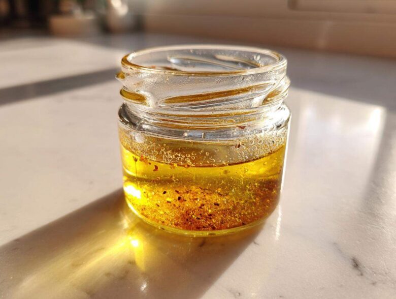 A small glass jar containing homemade salad dressing, featuring separated oil and spices, catching bright sunlight.