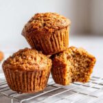 Three golden brown honey bran muffins stacked on a wire cooling rack, one muffin is broken open showing the texture.