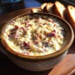 Close-up of a hot, cheesy reuben dip baked in a ceramic crock, served with toasted bread slices.