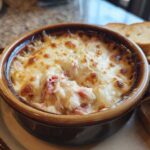 Close-up of a hot, cheesy reuben dip baked in a brown crock, served with toasted bread slices.