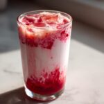 A tall glass filled with layered iced strawberry milk showing vibrant red syrup mixing with white milk and ice cubes.