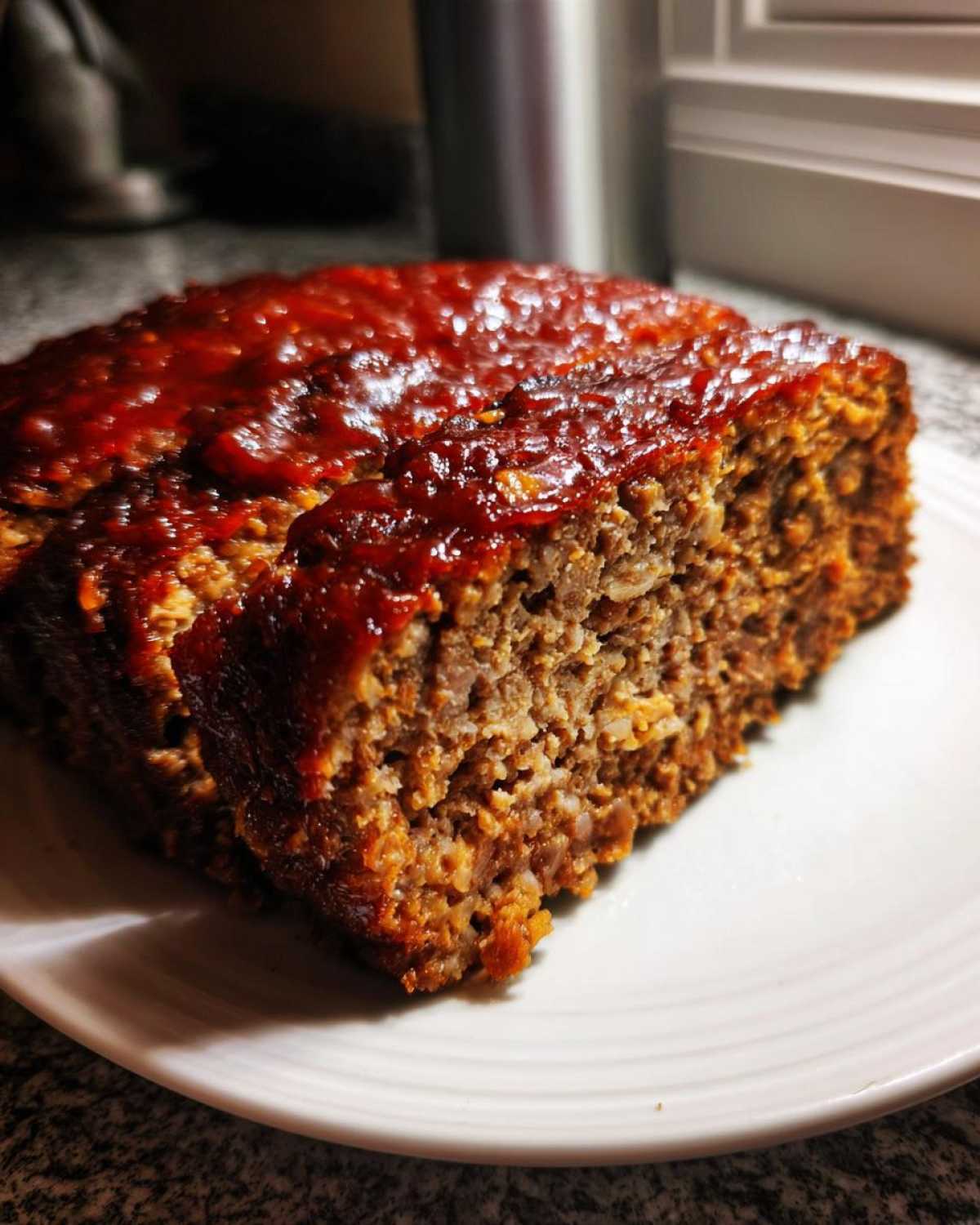 A close-up of a thick slice of juicy meatloaf topped with a shiny, dark red glaze, resting on a white plate.