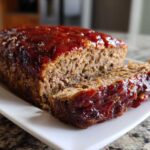 Close-up of a juicy meatloaf loaf topped with a thick, glossy red glaze, with one slice partially cut.