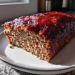 A thick slice of homemade meatloaf, topped with a shiny, thick red glaze, sitting on a white plate.