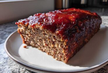 A thick slice of homemade meatloaf, topped with a shiny, thick red glaze, sitting on a white plate.