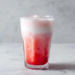 A tall glass filled with layered strawberry milk, showing a bright red base and a frothy white top layer.