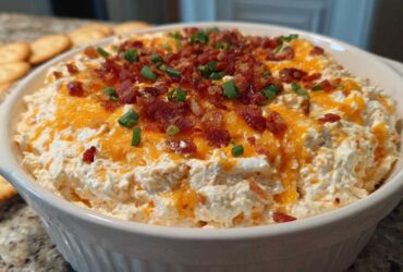 A close-up of creamy Million Dollar Dip topped with melted cheddar cheese, crispy bacon bits, and fresh chives.