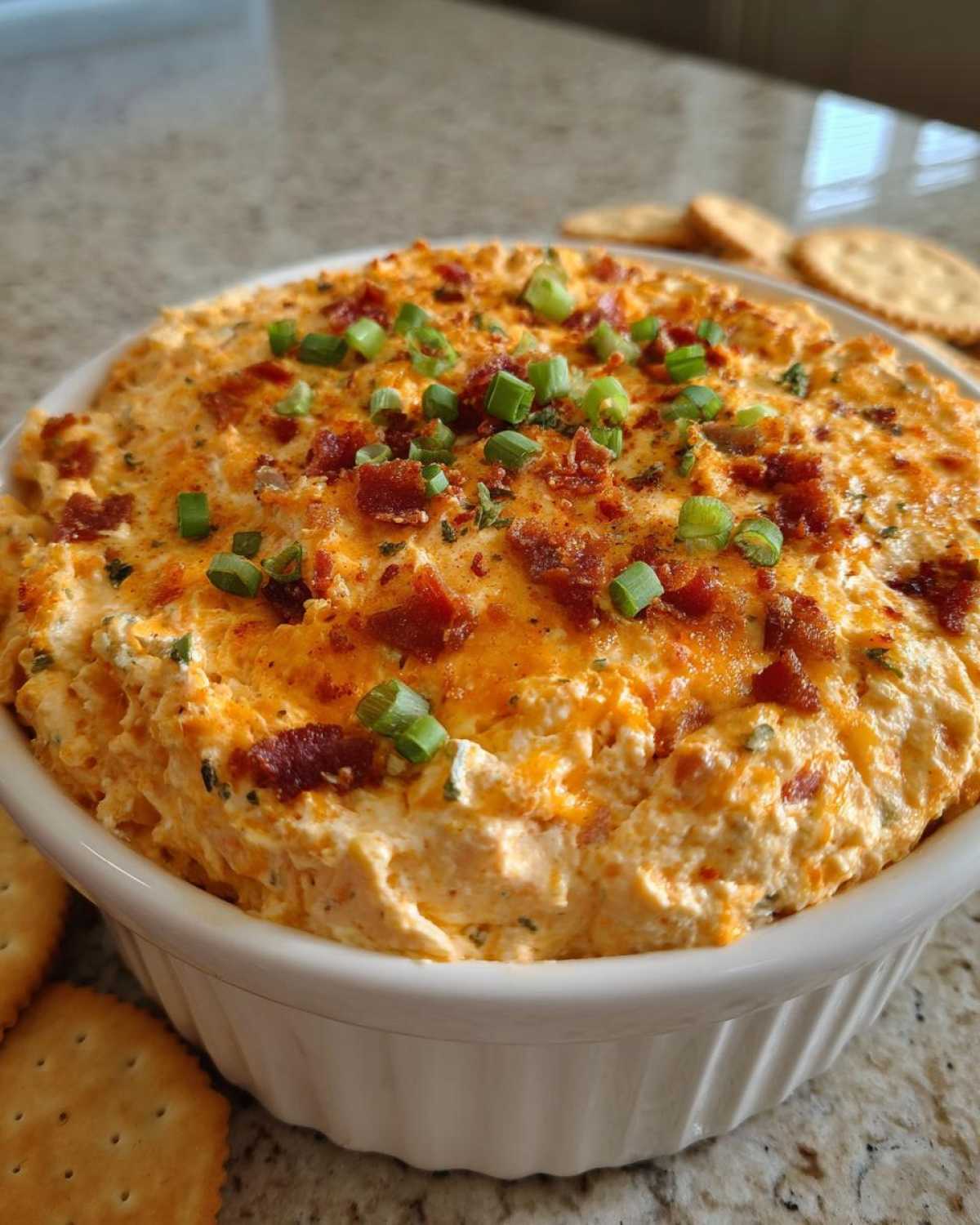 A close-up of creamy Million Dollar Dip served in a white bowl, topped with crumbled bacon and chopped green onions.