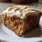 Close-up of a moist slice of applesauce cake topped with thick cream cheese frosting and a sprinkle of cinnamon.