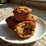 Close-up of three moist bran muffins, one cut open showing raisins inside, on a white plate.