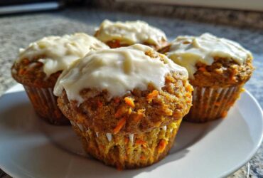 Four moist carrot cake muffins topped with thick cream cheese frosting, displayed on a white plate.