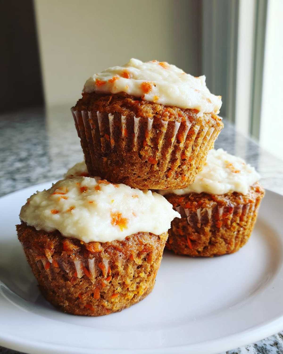 A stack of three moist carrot cake muffins topped with thick cream cheese frosting, sitting on a white plate.