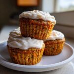 A stack of three moist carrot cake muffins topped with thick cream cheese frosting on a white plate.