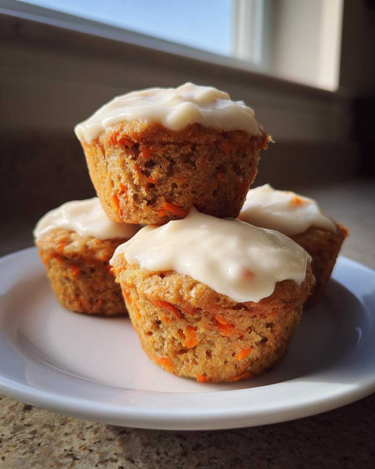 A stack of four moist carrot cake muffins topped with thick cream cheese frosting on a white plate.