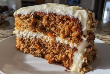 Close-up of a thick slice of moist carrot cake recipe with cream cheese frosting and chopped nuts.
