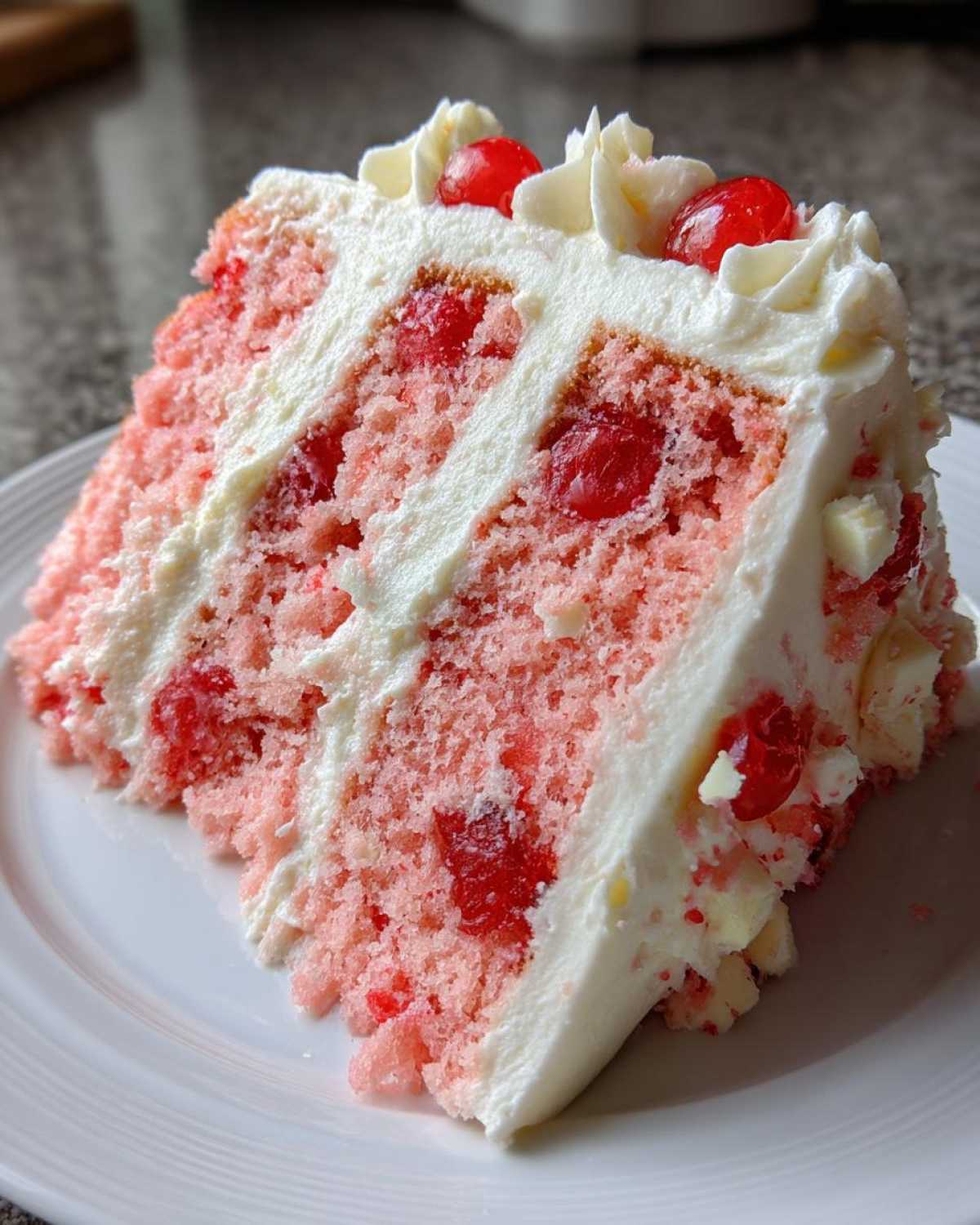 A close-up of a moist slice of pink cherry cake layered with white frosting and topped with maraschino cherries.
