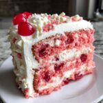 Close-up of a moist cherry cake slice showing pink layers, white frosting, and whole cherries.