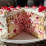 A slice removed from a moist cherry cake showing layers of yellow cake, pink cherry filling, and white frosting, topped with maraschino cherries.