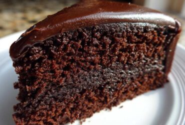 Close-up of a rich, moist slice of chocolate cake recipe topped with glossy dark chocolate frosting.