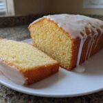 A loaf of moist lemon pound cake, partially sliced, topped with a thick, white lemon glaze dripping down the sides.