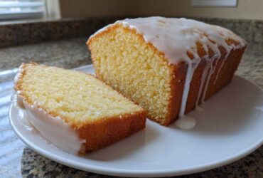 A loaf of moist lemon pound cake, partially sliced, topped with a thick, white lemon glaze dripping down the sides.