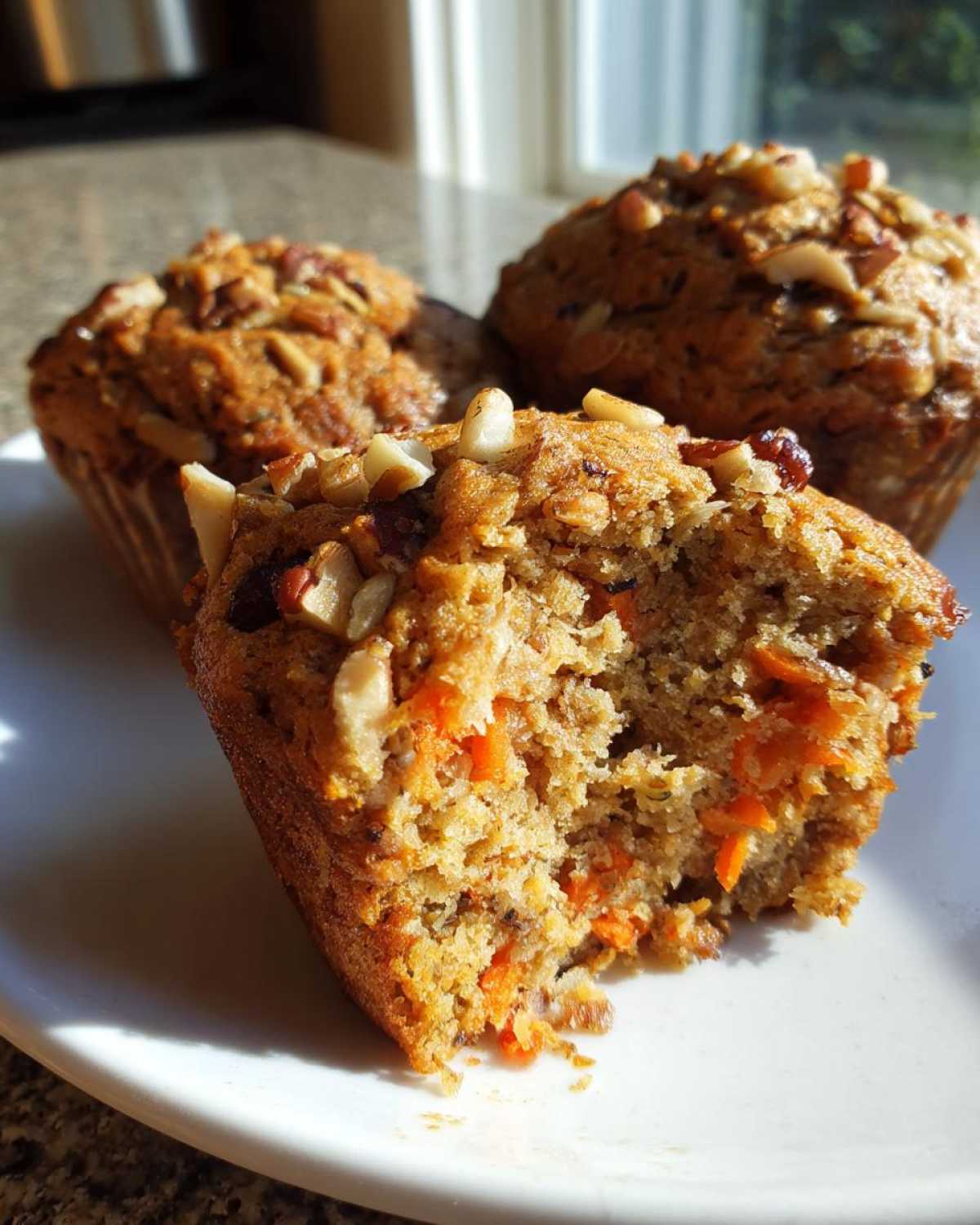 A close-up of a morning glory muffin broken open, showing shredded carrots and nuts inside, with two more muffins behind it.