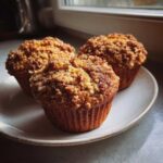 Three freshly baked oatmeal muffins with a thick, golden-brown streusel topping resting on a light-colored plate.