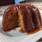 A moist, dark brown rum cake with a slice cut out, sitting on a white plate covered in a shiny rum glaze.