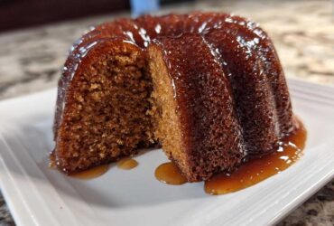 A moist, dark brown rum cake with a slice cut out, sitting on a white plate covered in a shiny rum glaze.