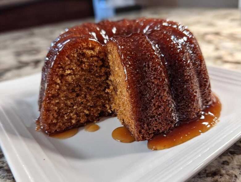 A moist, dark brown rum cake with a slice cut out, sitting on a white plate covered in a shiny rum glaze.