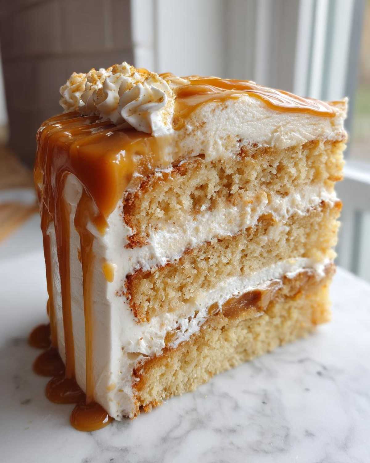 A tall, moist slice of caramel cake featuring three layers of sponge, white frosting, and a generous caramel drizzle.
