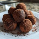 A stack of homemade, dark chocolate protein balls heavily dusted with cocoa powder, served on a white plate.