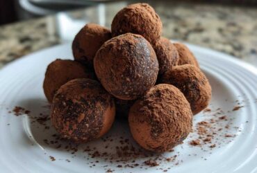 A stack of homemade, dark chocolate protein balls heavily dusted with cocoa powder, served on a white plate.