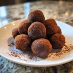 A stack of homemade, dark chocolate protein balls generously dusted with cocoa powder on a white plate.