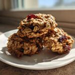 Three healthy oatmeal breakfast cookie treats stacked on a white plate, featuring visible oats and dried cranberries.