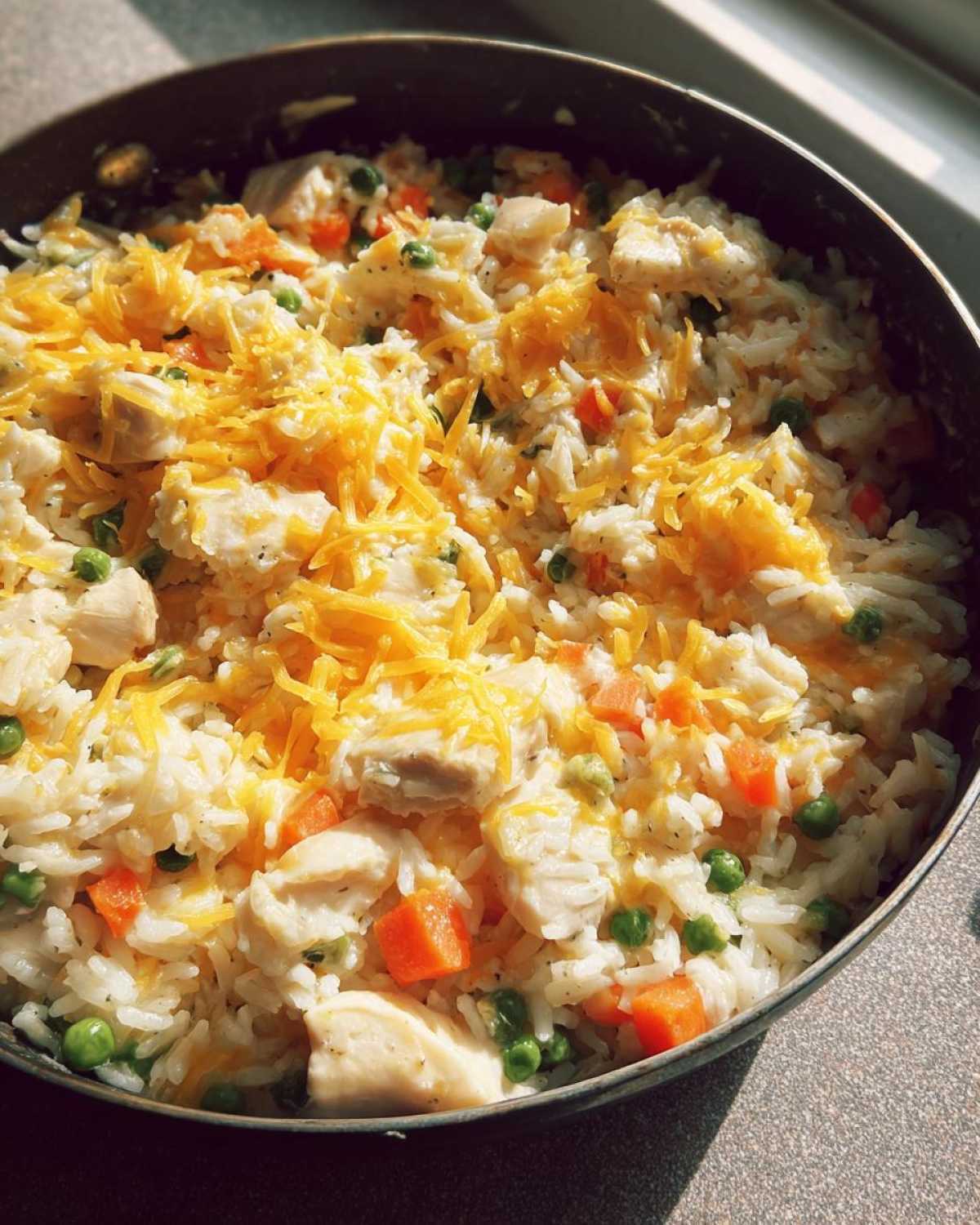 Close-up of a skillet filled with creamy chicken and rice mixed with peas and carrots, topped with shredded cheddar cheese.