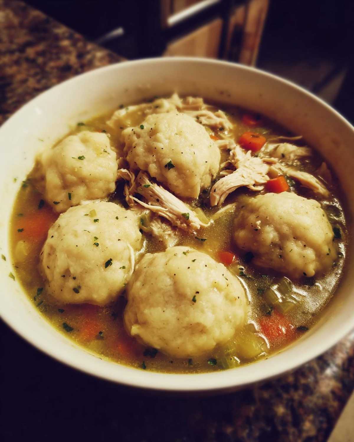 A close-up of a white bowl filled with rich broth, shredded chicken, carrots, and four large, fluffy chicken and dumpling pieces.
