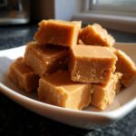 A stack of golden brown, melt-in-mouth penuche fudge squares piled on a white dish.