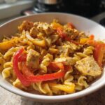 A close-up of a white bowl filled with creamy Rasta pasta, featuring chunks of seasoned chicken and vibrant red and yellow bell peppers.