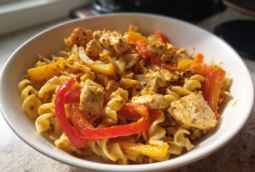 A close-up of a white bowl filled with creamy Rasta pasta, featuring chunks of seasoned chicken and vibrant red and yellow bell peppers.