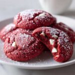 A stack of soft red velvet cake mix cookies dusted with powdered sugar, one cookie broken open showing white chocolate chips.