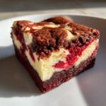 A close-up of a single, rich red velvet cheesecake brownie square on a white plate.