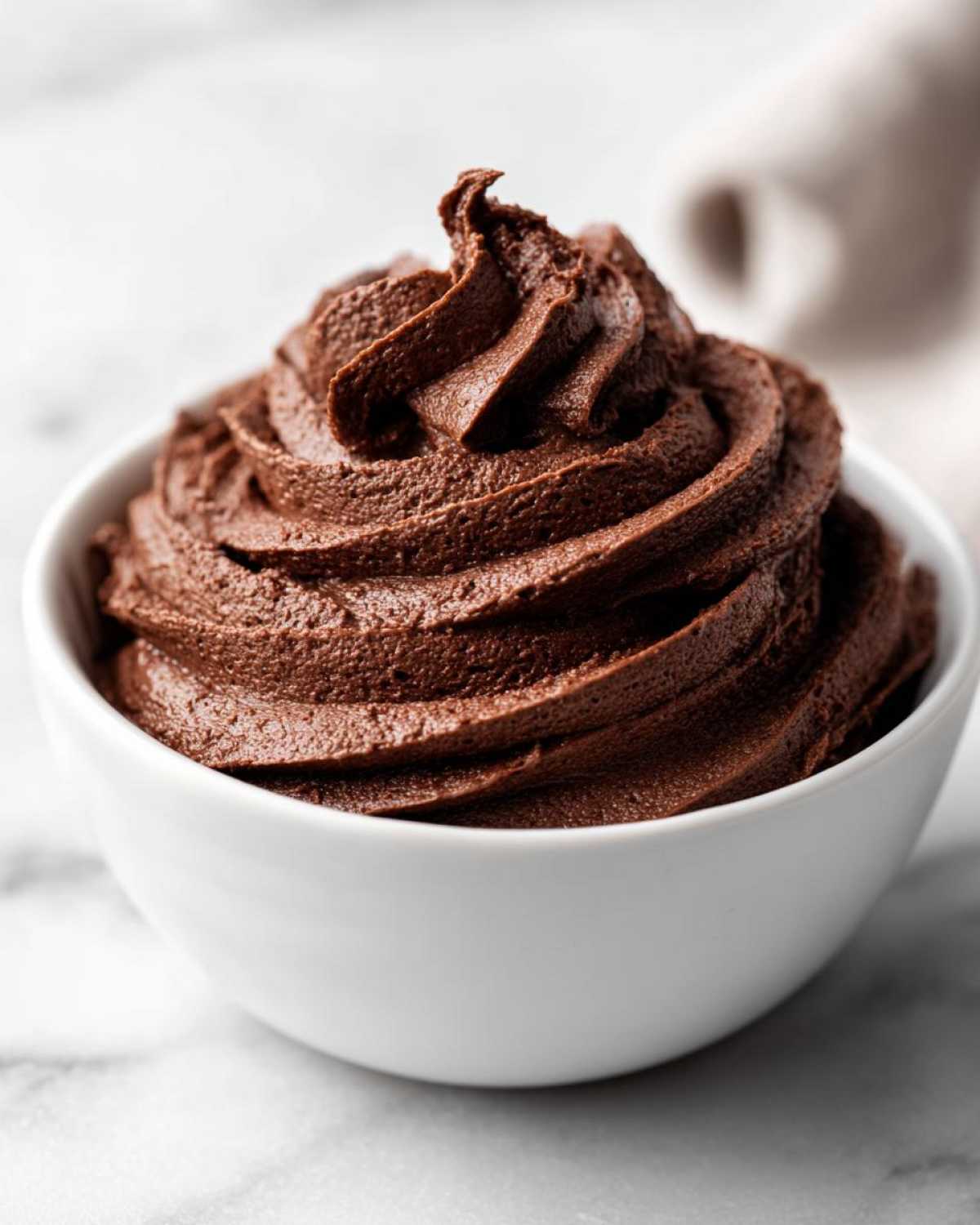 A close-up shot of rich, dark chocolate frosting swirled high in a small white bowl.