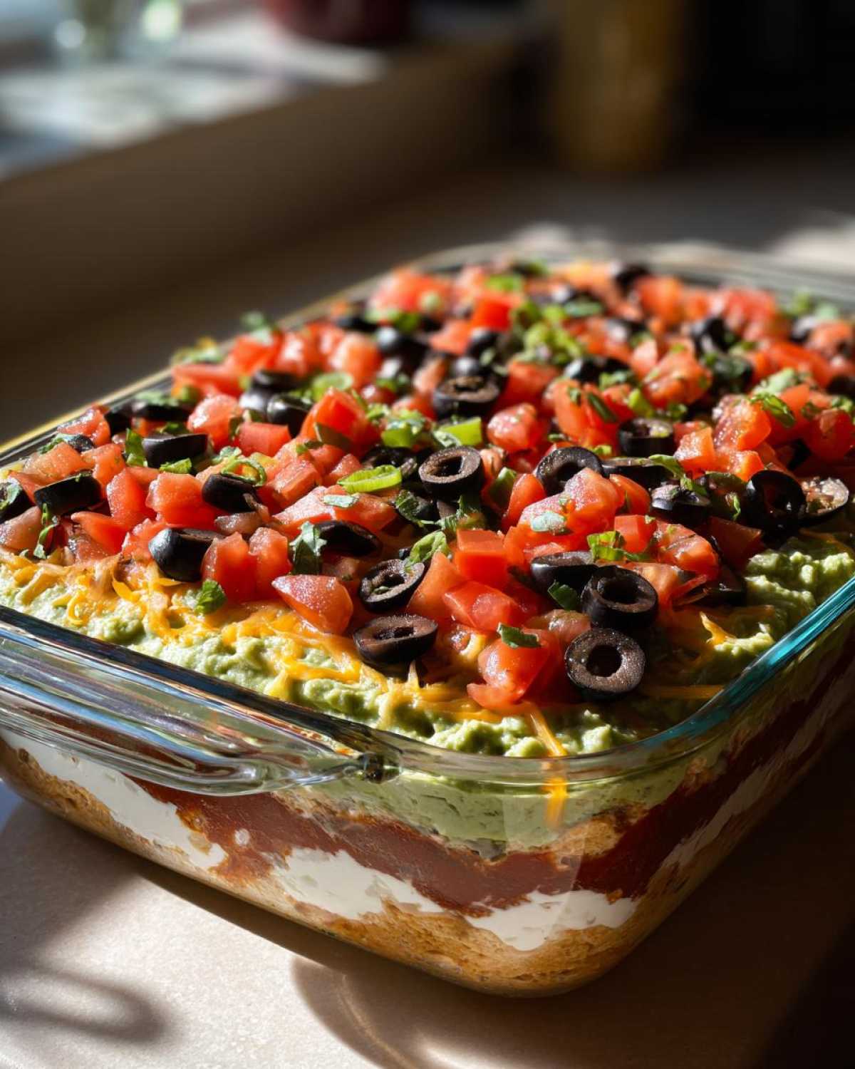 A clear glass dish filled with a vibrant Seven layer dip, topped with guacamole, cheese, tomatoes, and olives.