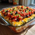 A vibrant, fully assembled Seven layer dip in a clear glass baking dish, topped with tomatoes, olives, and green onions.