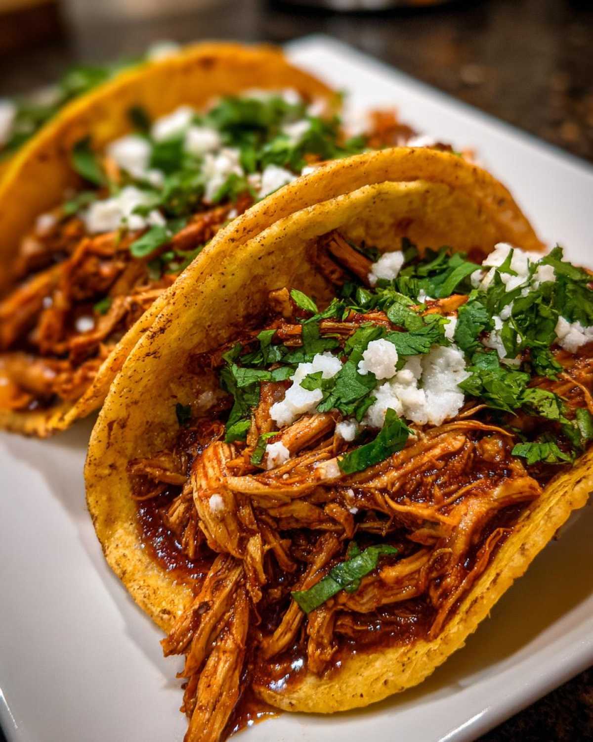 Close-up of two delicious chicken tacos filled with saucy shredded meat, topped with cotija cheese and cilantro.